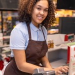 Happy young mixed-race cashier with dark wavy hair pressing button on cashbox