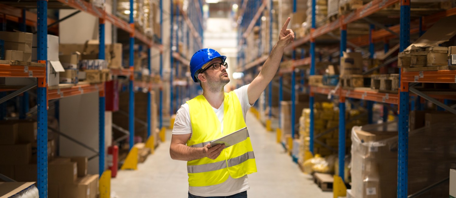 Warehouse worker in protective reflective uniform with hardhat checking inventory and counting product on shelf in large storage area.