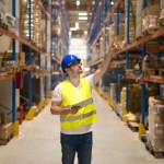 Warehouse worker in protective reflective uniform with hardhat checking inventory and counting product on shelf in large storage area.