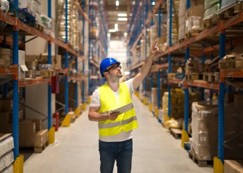Warehouse worker in protective reflective uniform with hardhat checking inventory and counting product on shelf in large storage area.