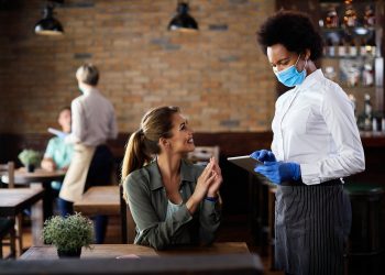 Happy black waitress wearing protective face mask while taking o